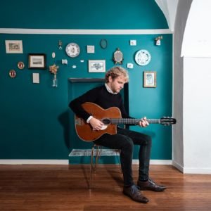 Fanou Torracinta assis sur une chaise devant un mur bleu en train de jouer de la guitare.
