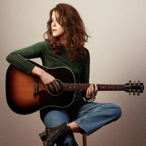 Jeanne assise sur un tabouret avec sa guitare, habillée en jean et pull vert. 